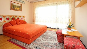 Ein Schlafzimmer mit einem großen Bett, einem kleinen Tisch und einem Sessel. Ein Fenster mit Vorhängen, ein Bild an der Wand und ein Teppich mit Muster.