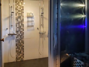A bathroom with a shower, a toilet, and a sink. There is a tiled wall with a checkered pattern and a shelf with toiletries. - Aarrekammi by Interhome (Inari)