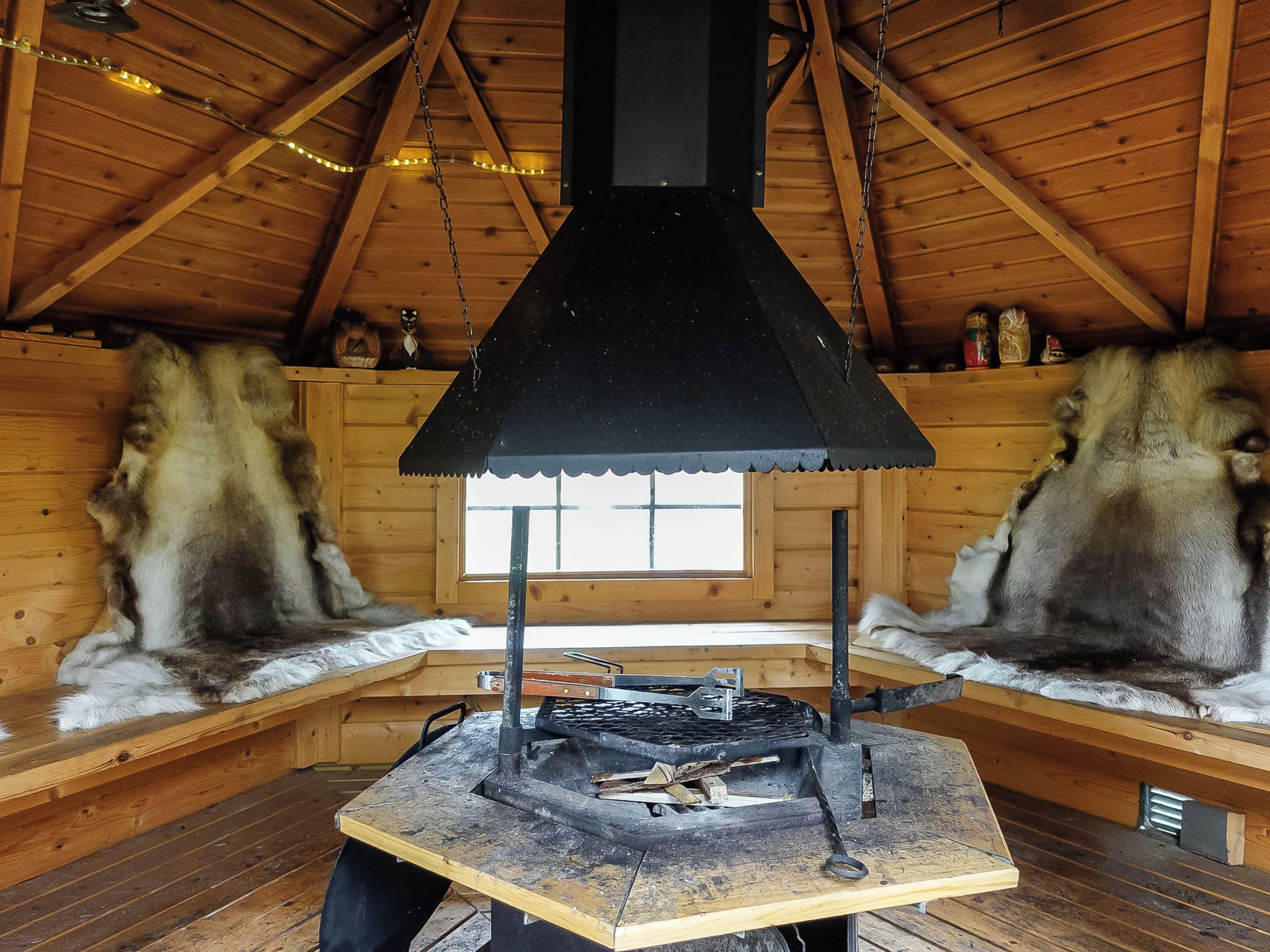 A rustic cabin interior with a central wood-burning stove, wooden benches, and a sloped ceiling.