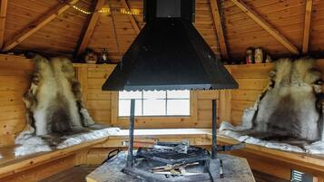 A rustic cabin interior with a central wood-burning stove, wooden benches, and a sloped ceiling.