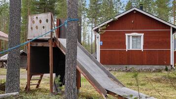 A wooden play structure with a slide, a red building, and a forested background.