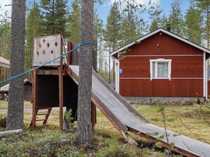 A wooden play structure with a slide, a red building, and a forested background.