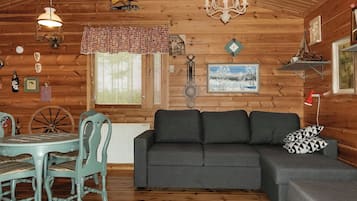 Ein gemütliches Blockhauss-Interieur mit Holzbalkendecke, einem Esstisch mit Stühlen, einer Couch und einer Kandelaberleuchte.