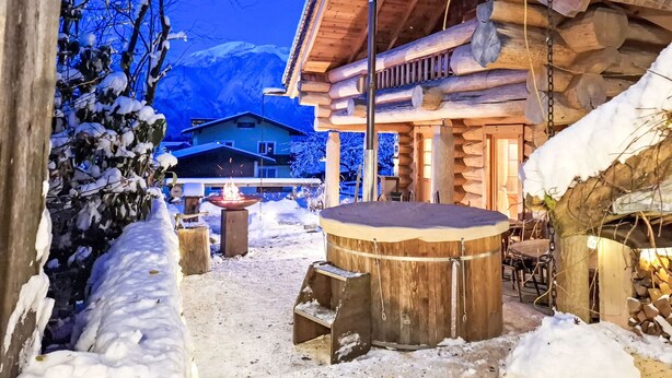 Una cabaña de troncos con fachada de madera, una fogata en el centro y un paisaje cubierto de nieve.