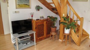 A living room with a wooden staircase, a flat-screen TV on a stand, and several potted plants.