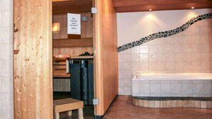 A sauna room with a wooden bench, a bench with a towel, a black heater, and a tiled wall with a mosaic border.