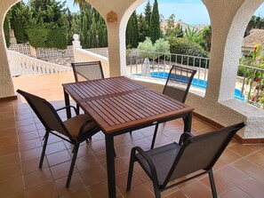 Eine überdachte Terrasse mit Holztisch und -stühlen, ein Pool im Hintergrund und Blick auf Grünflächen und Gebäude.