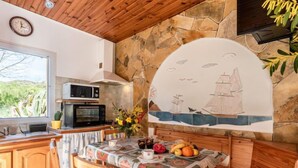 Une cuisine rustique avec des meubles en bois, un mur en pierre, une fresque représentant des navires et une table à manger agrémentée d’un panier de fruits.