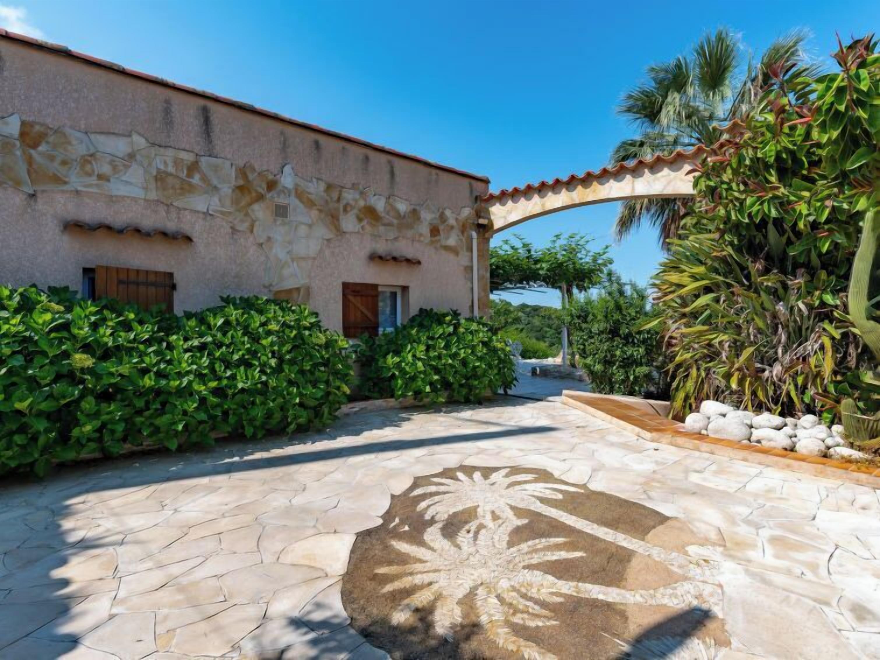 Un bâtiment doté d’une façade en pierre, d’un patio carrelé orné d’un motif de palmiers et d’une végétation luxuriante.