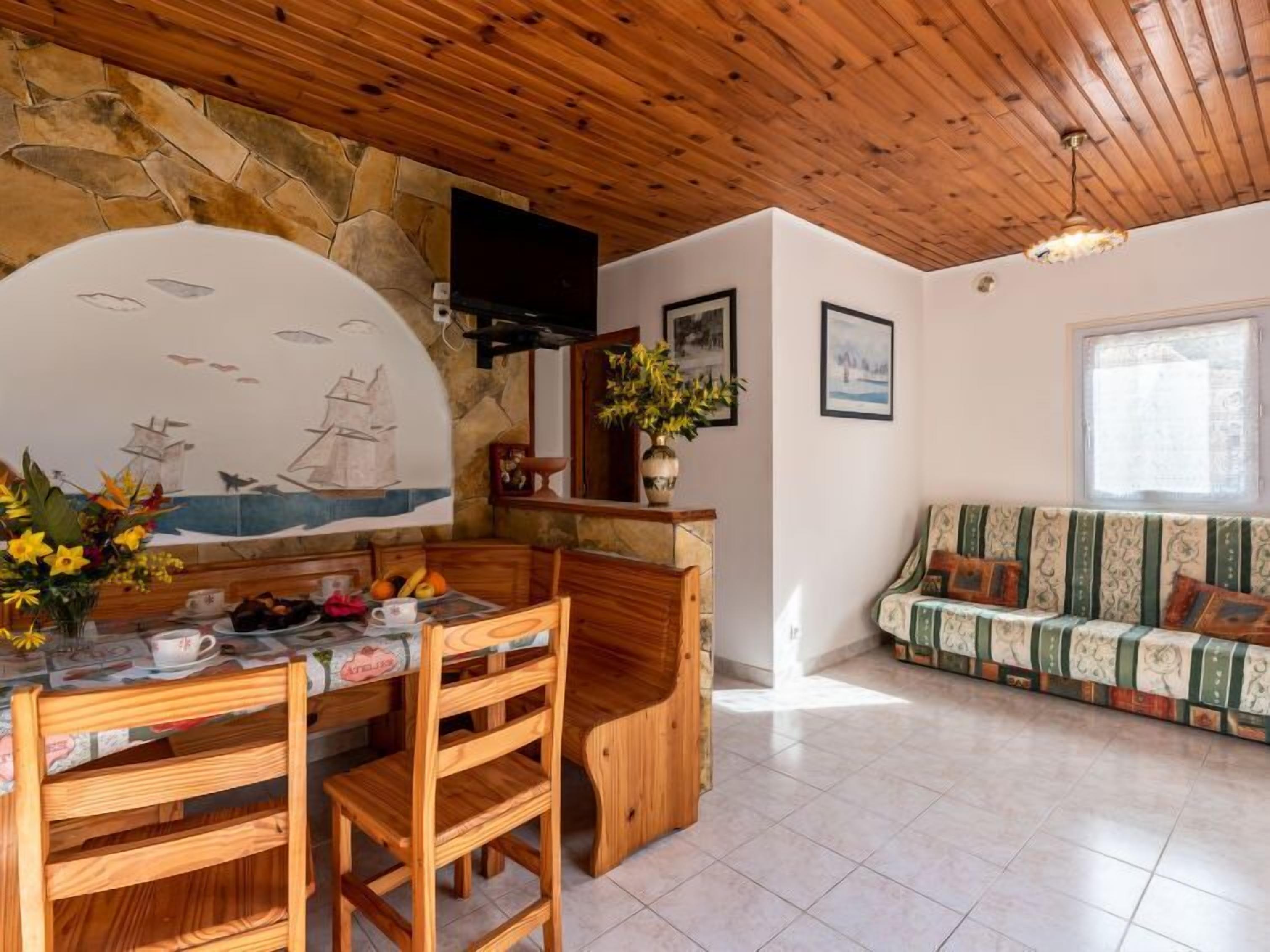Ein gemütliches Zimmer mit Steinwand, Holzbalkendecke, einem Esstisch mit Stühlen, einer Couch und einem an der Wand befestigten Fernseher.