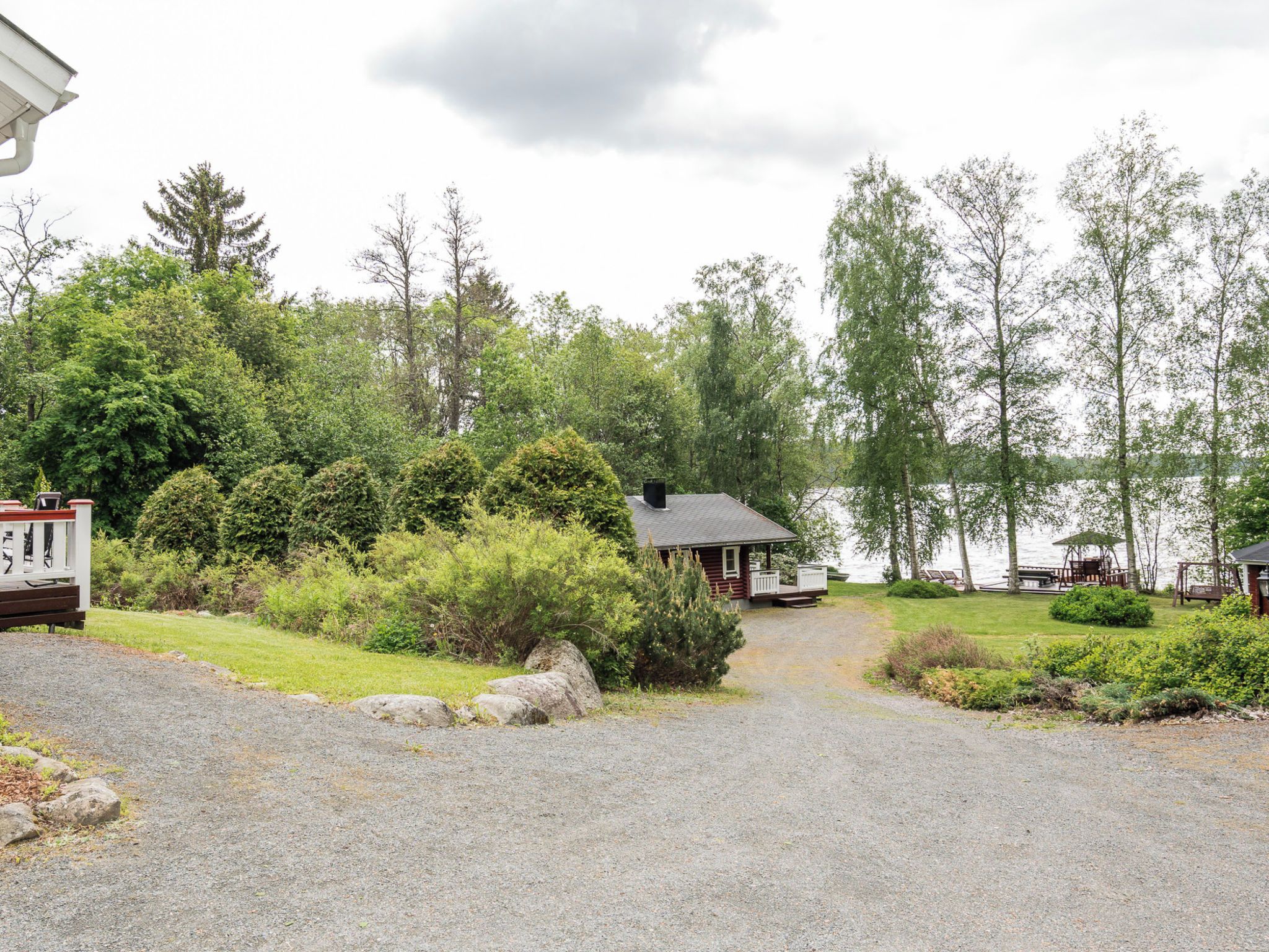 A gravel driveway leading to a cabin with a small porch, surrounded by dense greenery and a lake.