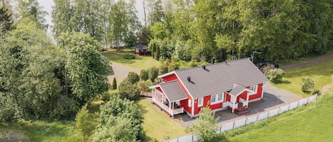 Ett rött hus med en veranda, omgivet av ett vitt staket, nära en sjö med träd.