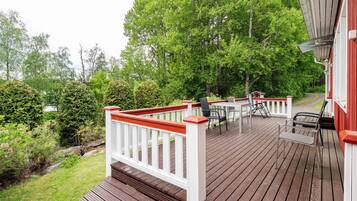 Une terrasse en bois avec des meubles d’extérieur, une balustrade blanche et un bâtiment rouge en arrière-plan.
