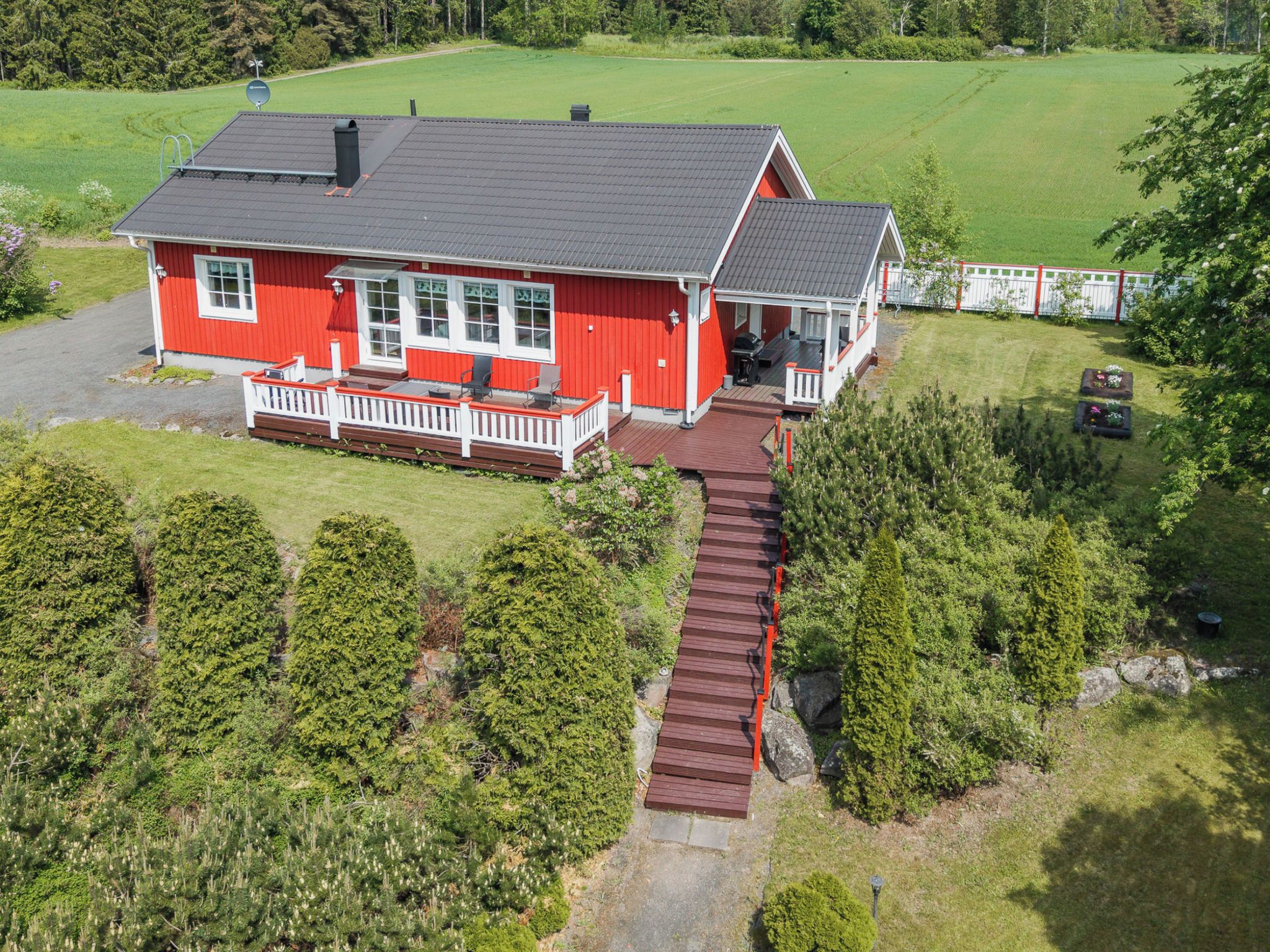 Une maison rouge avec un porche et une terrasse, entourée d’une pelouse bien entretenue et d’arbres.