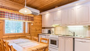 A wooden cabin interior with a dining table, chairs, a kitchen with white appliances, and a window with a plaid curtain.