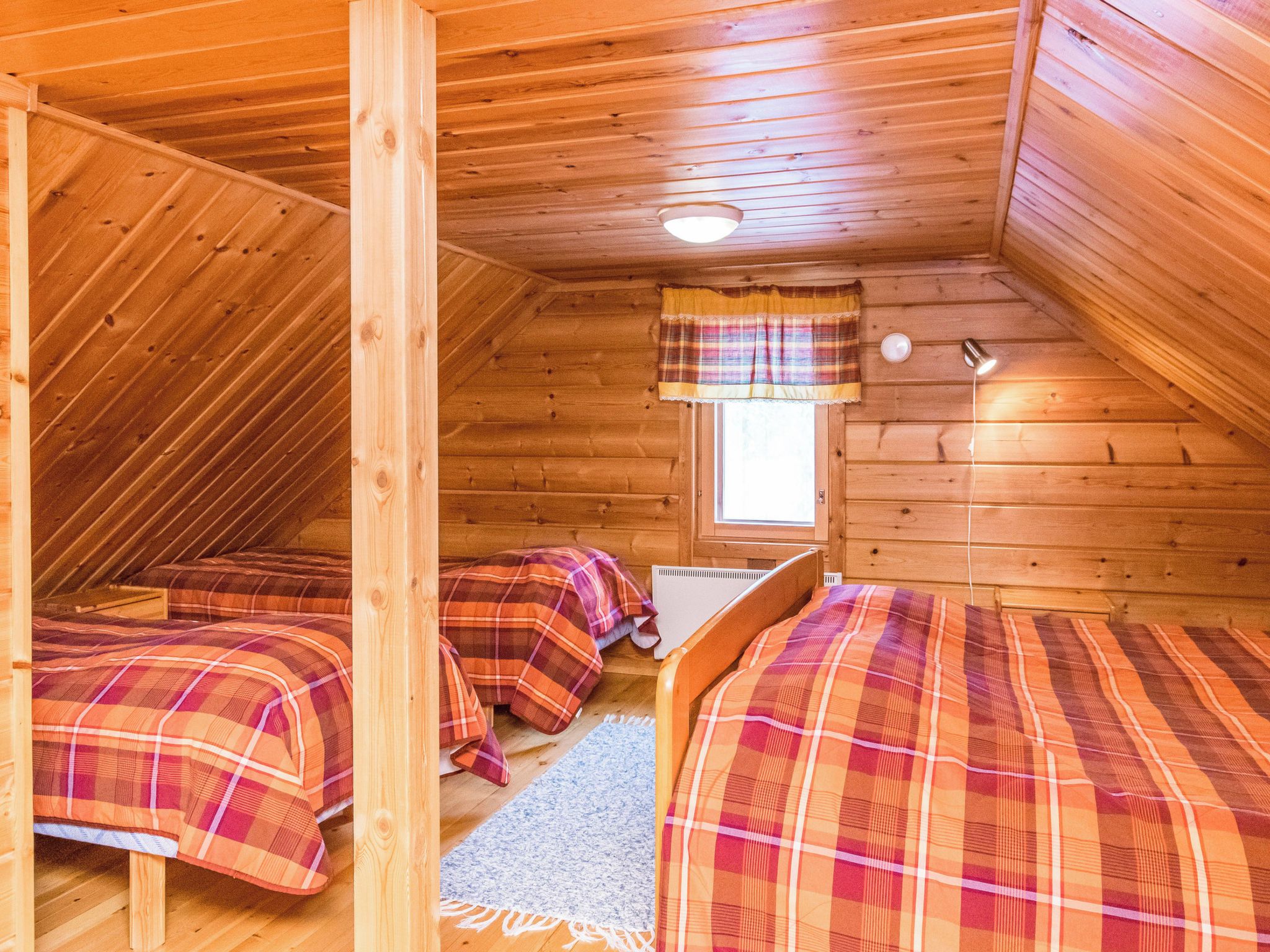 Een knus slaapkamer onder het dak, met houten wanden en schuine plafonds. Er staan twee bedden met geruite beddengoed op en er is een klein raam met gordijn.