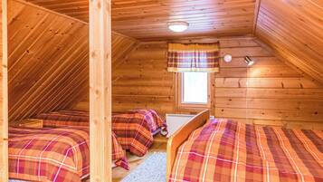A cozy attic bedroom with wooden walls and sloped ceilings. There are two beds with plaid bedding and a small window with a curtain.