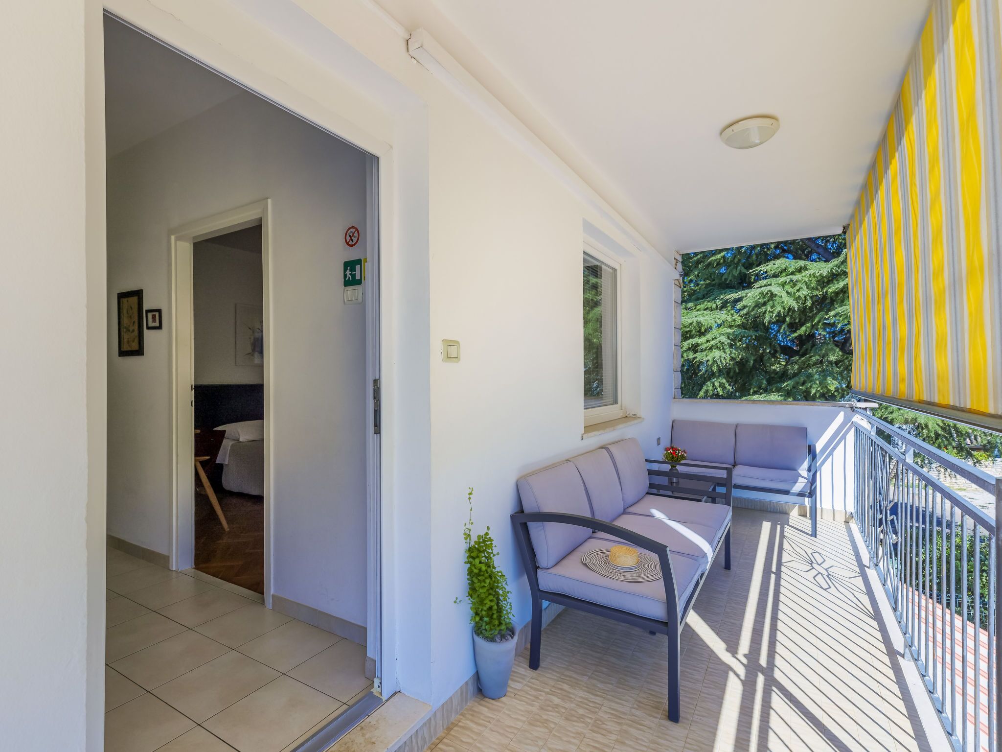 Een balkon met een bank en een bloempot, een deur met een groen vertrekbord, en een raam met uitzicht op bomen.