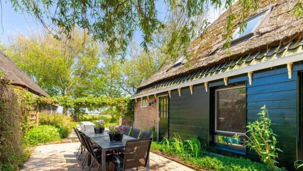 Une terrasse avec une table et des chaises, entourée de verdure et bordée d’un bâtiment au toit de chaume.