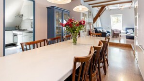 Un coin repas avec une longue table, des chaises en bois et des lampes suspendues. On y trouve un buffet bleu et un vase avec des fleurs sur la table.