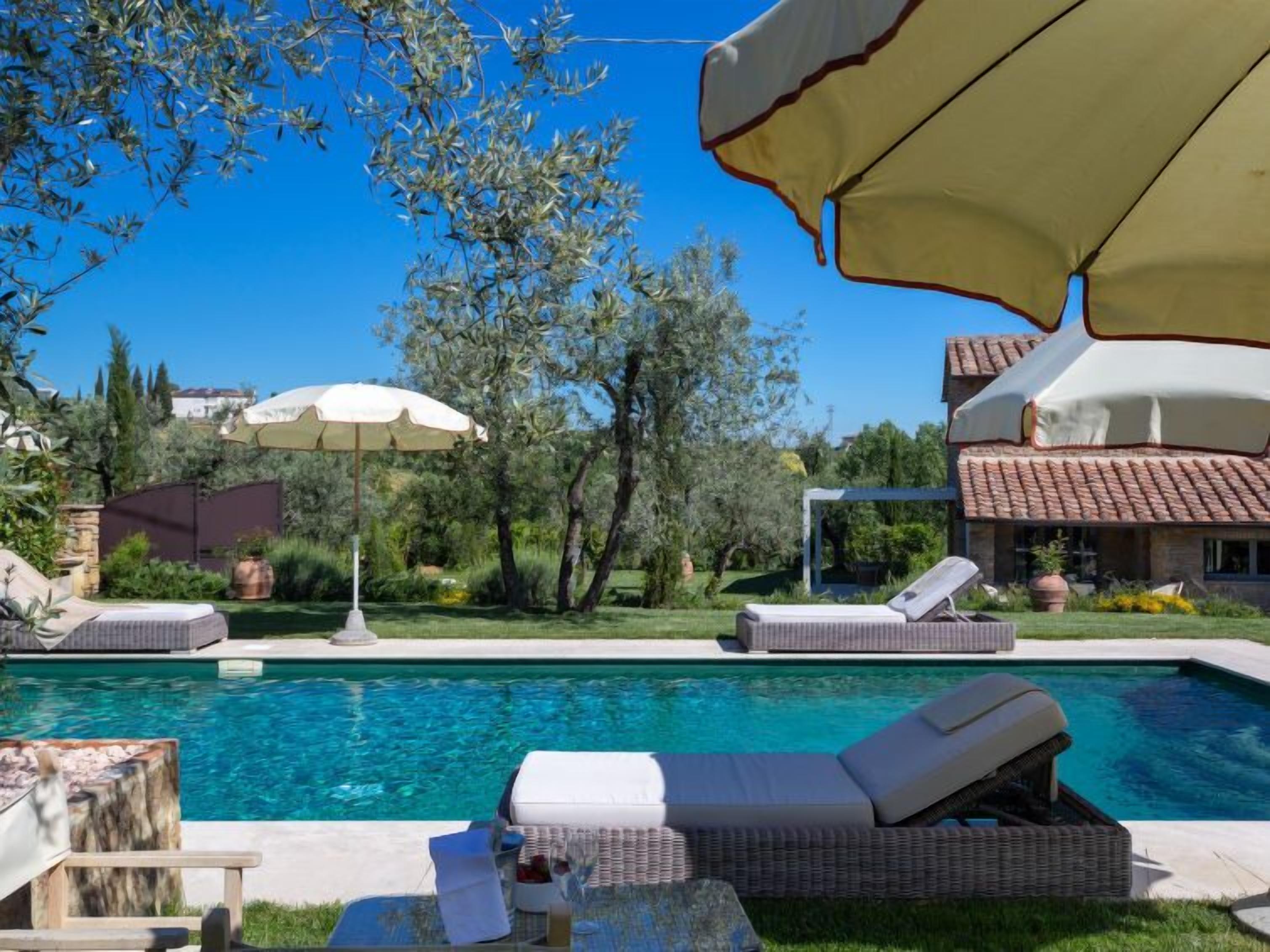 Ein Swimmingpool mit Liegestühlen und Sonnenschirmen, umgeben von Grünflächen und einem Haus mit Ziegeldach.