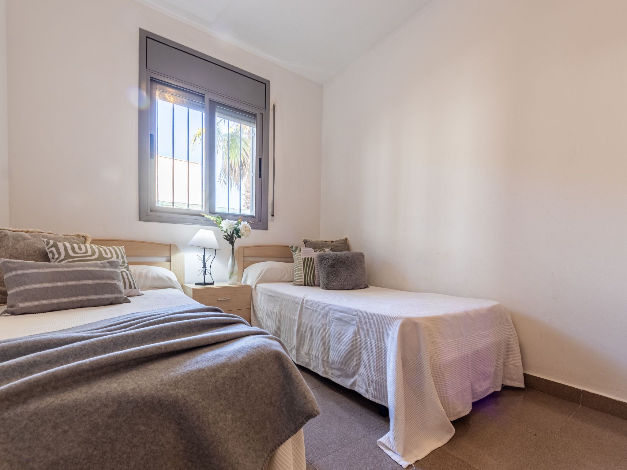A bedroom with two beds, a window with blinds, a bedside table with a lamp, and a vase with flowers.
