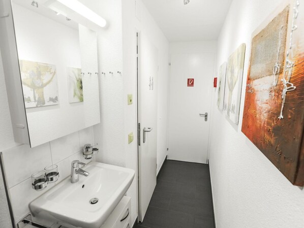 Ein schmaler Flur mit einem weißen Waschbecken und Spiegel, einer Tür mit einem roten Schild und Kunstwerken an den Wänden.