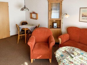 Une pièce chaleureuse avec deux fauteuils orange, une petite table recouverte d’une nappe à motifs floraux, un buffet en bois aux portes vitrées, une lampe suspendue et un tableau encadré au mur.