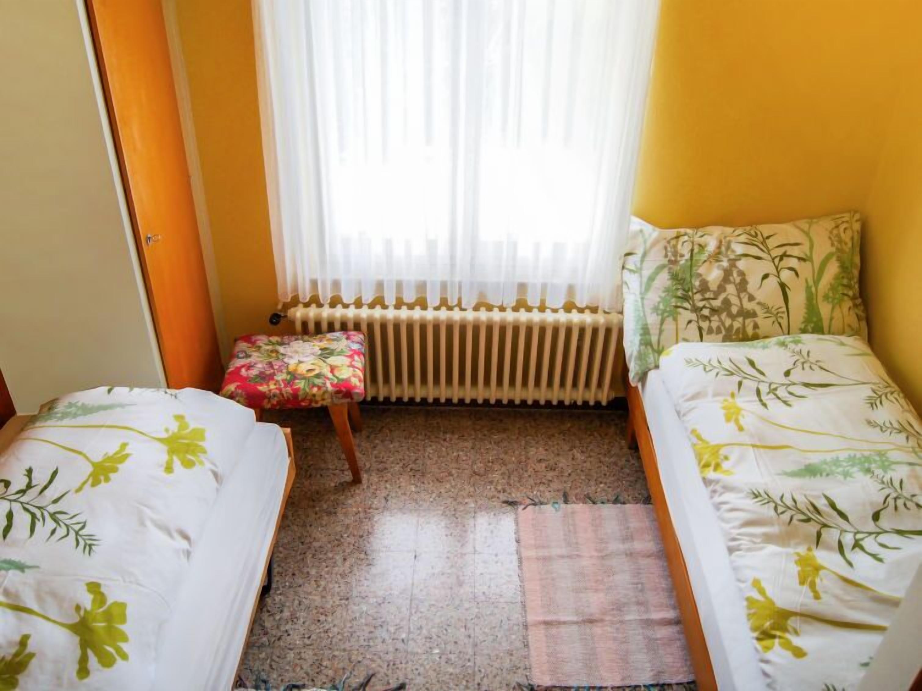A small, cozy room with two single beds, a radiator, a window with sheer curtains, and a small stool.