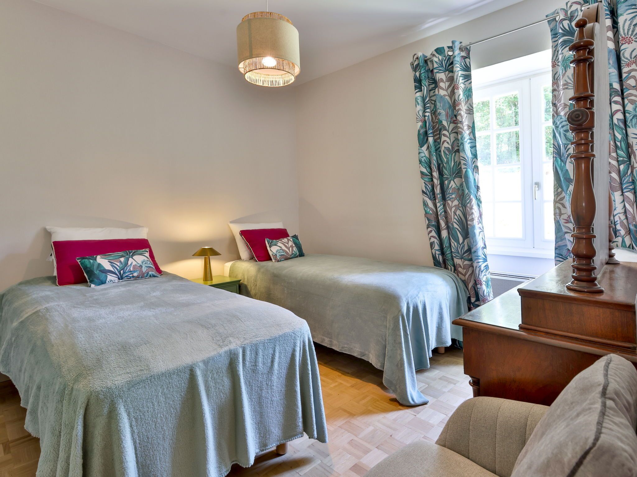 A bedroom with two beds, a nightstand, a lamp, a wooden dresser, and a window with curtains.
