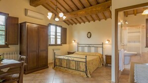 Une chambre à coucher avec un lit, des poutres en bois au plafond, une fenêtre, un radiateur et une salle de bain visible à travers une porte ouverte.