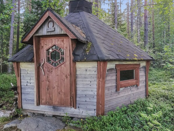 A small wooden cabin with a hexagonal door and a window, surrounded by a forest.