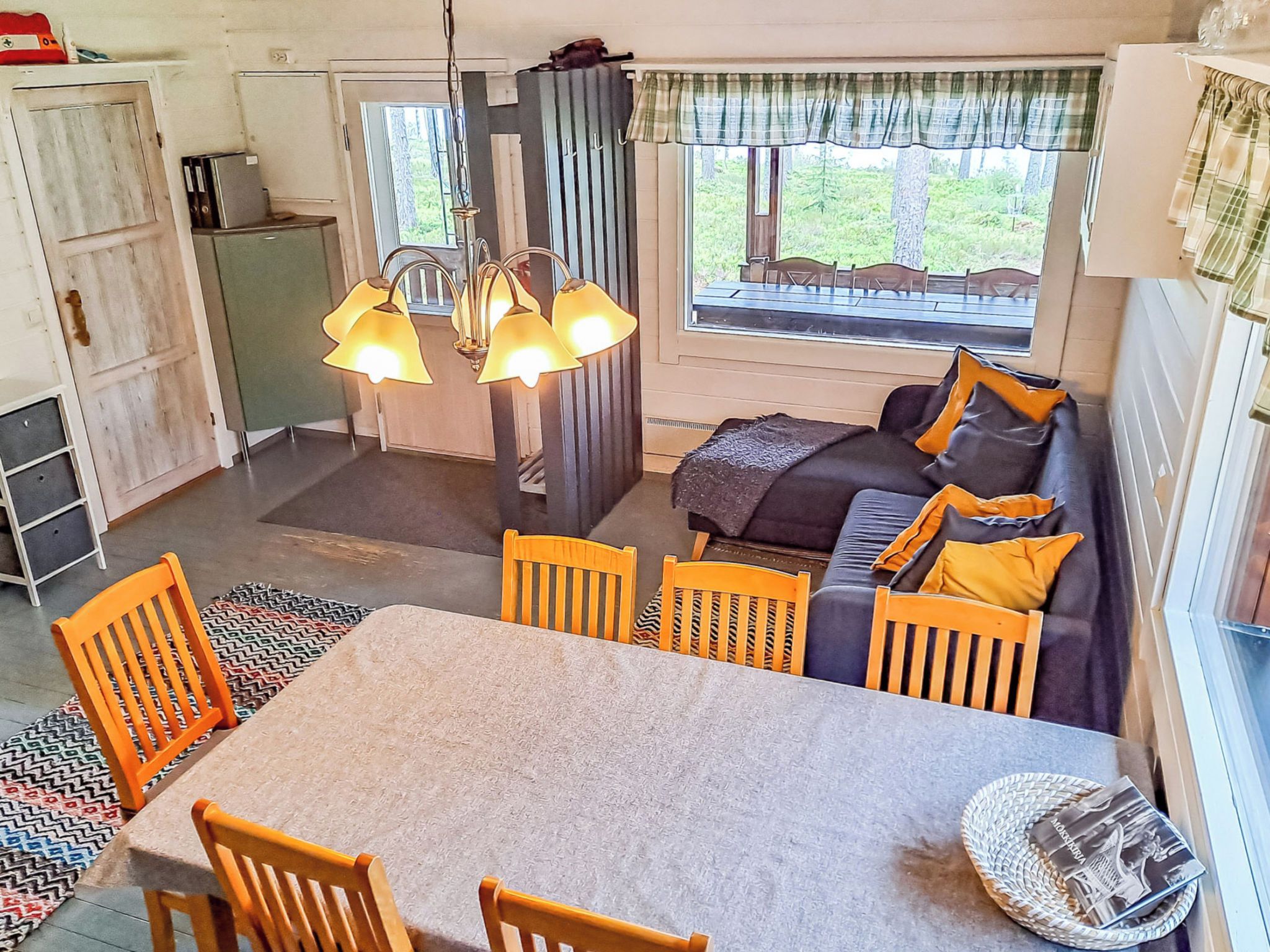 Een gezellig interieur met een eettafel, stoelen, een bank met kussens, een hanglamp en uitzicht door een raam.