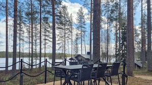 A wooden deck with a glass table and chairs overlooking a lake, surrounded by tall trees. - Pilkkaniemi by Interhome (Juuka)
