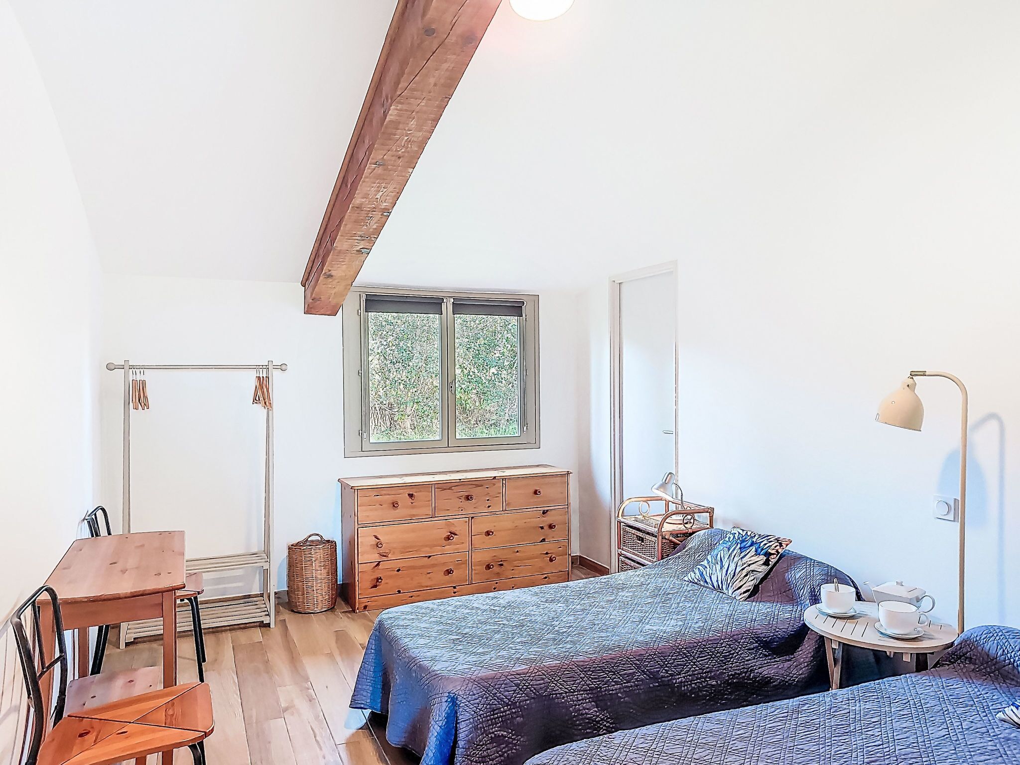 Un dormitorio con un aparador de madera, una cama con ropa de cama estampada, una mesita con tazas y una lámpara, y una ventana con vista a la vegetación.