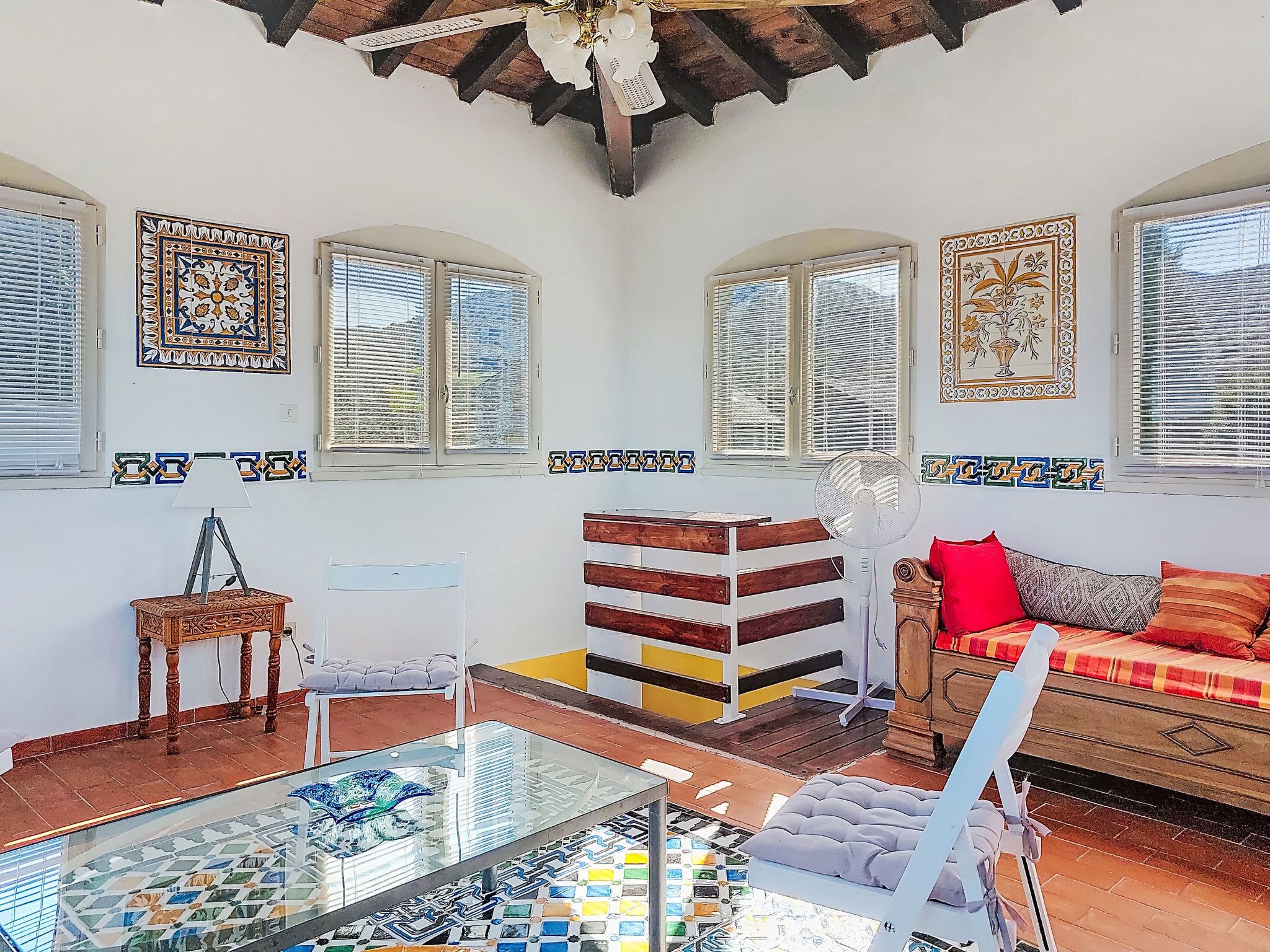 A room with a glass coffee table, a wooden bench, a sofa with cushions, and a fan.