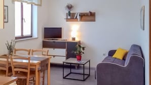 Un salon moderne avec un canapé gris, une table à manger en bois et des chaises. On y trouve un téléviseur posé sur un meuble, une petite table avec une plante et une étagère ornée d’objets décoratifs.