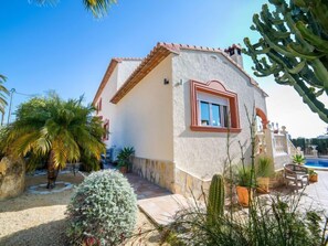 Una casa de dos pisos con techo de tejas rojas, una ventana pequeña con marco blanco, y un área de piscina con una silla y una mesa.