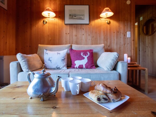 A cozy room with a wooden wall, a sofa with patterned cushions, a table with a teapot, cups, and pastries. - Chalet Lorila by Interhome (Villars)