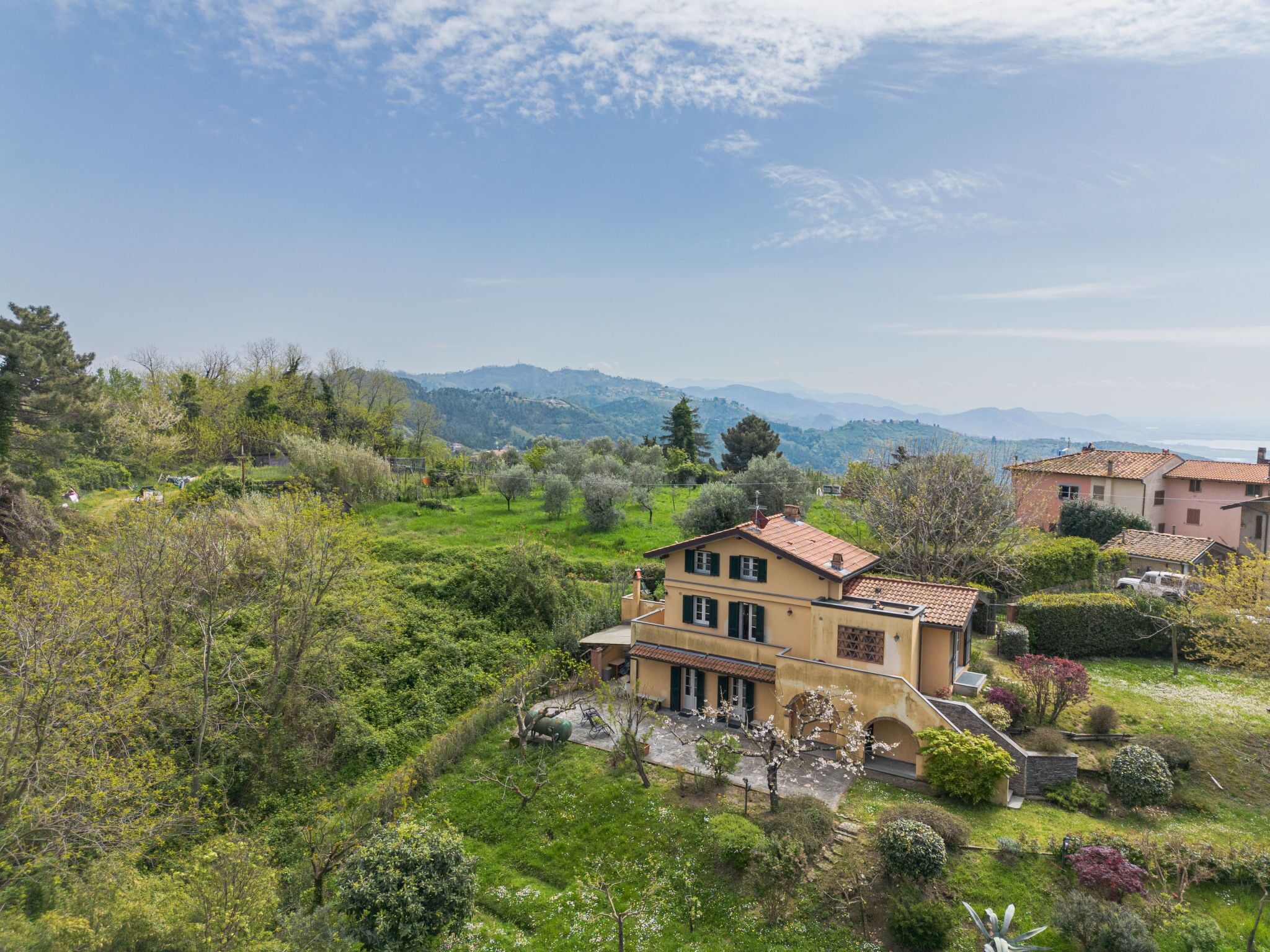 Una villa su una collina con un giardino e vista sulle montagne, sotto un cielo sereno.