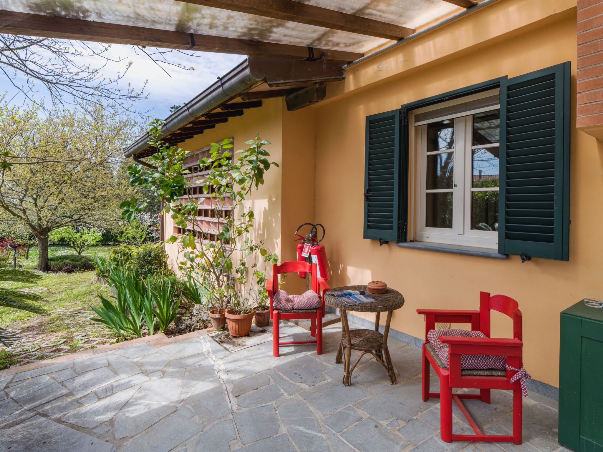 Eine überdachte Terrasse mit Steinboden, zwei roten Stühlen, einem Korbstuhl-Tisch und einem Fenster mit grünen Rollläden.