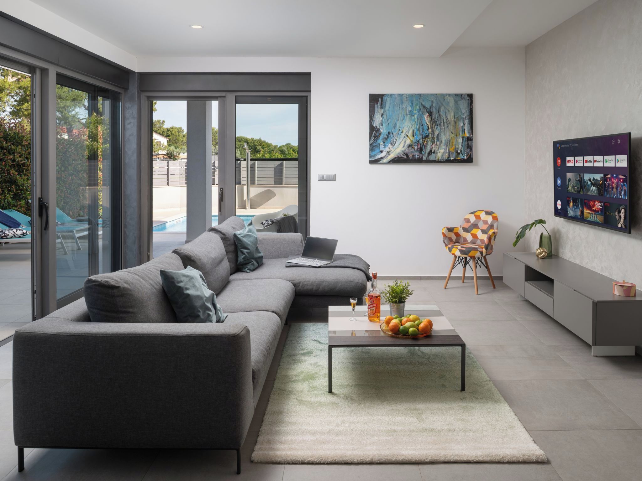 A modern living room with a grey sectional sofa, a coffee table with fruits and a bottle, a TV mounted on the wall, and a large window overlooking a pool.