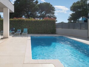 Una piscina con agua cristalina azul, rodeada por una solana con baldosas y un seto.