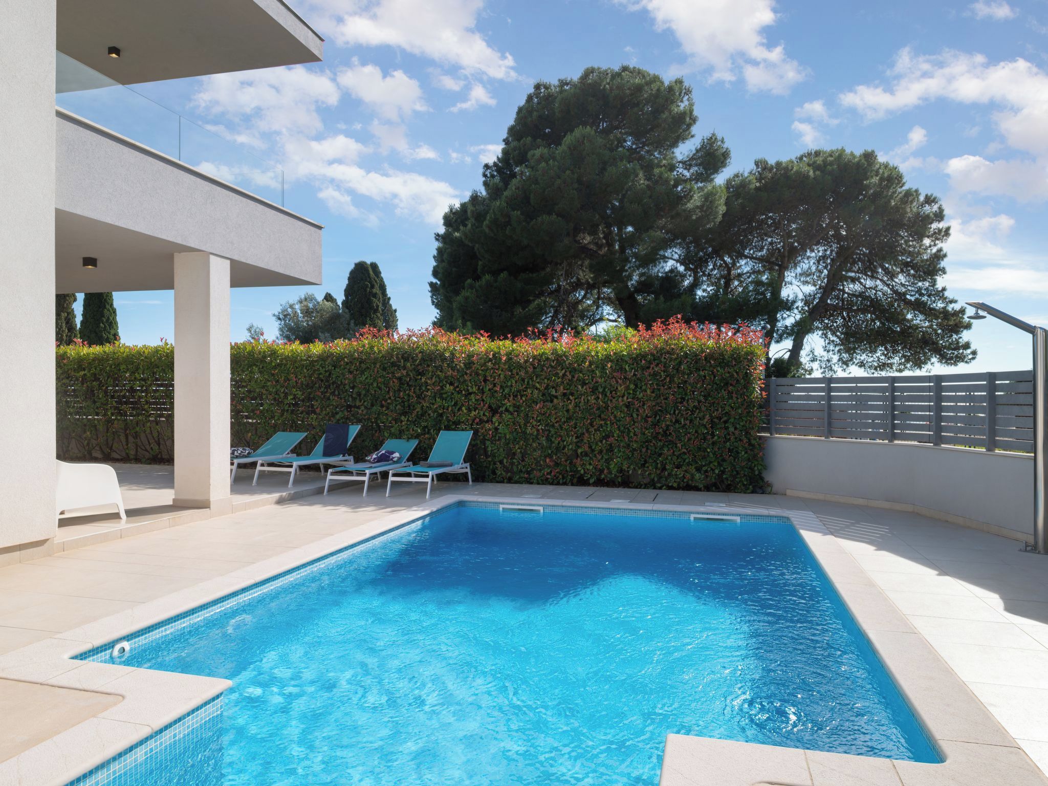 A rectangular swimming pool with clear blue water, surrounded by a tiled deck. There are lounge chairs and a hedge with red flowers.