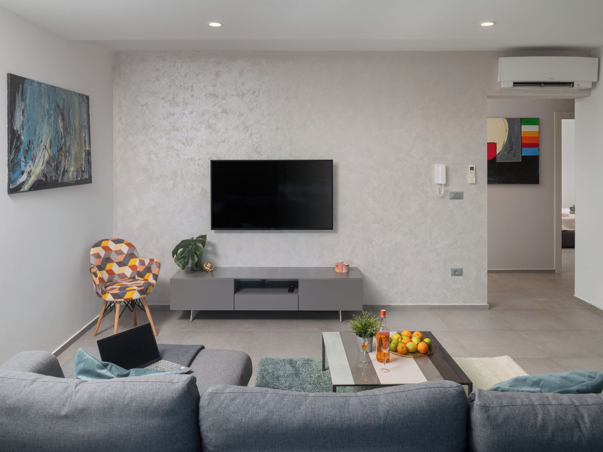A modern living room with a flat-screen TV mounted on a textured wall, a gray sofa, a coffee table with fruits and drinks, and a colorful chair.