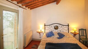 A bedroom with a wooden ceiling, a bed with a metal headboard, two bedside lamps, a chair, and a window with blinds. - Le Bonatte by Interhome (Radda in Chianti)