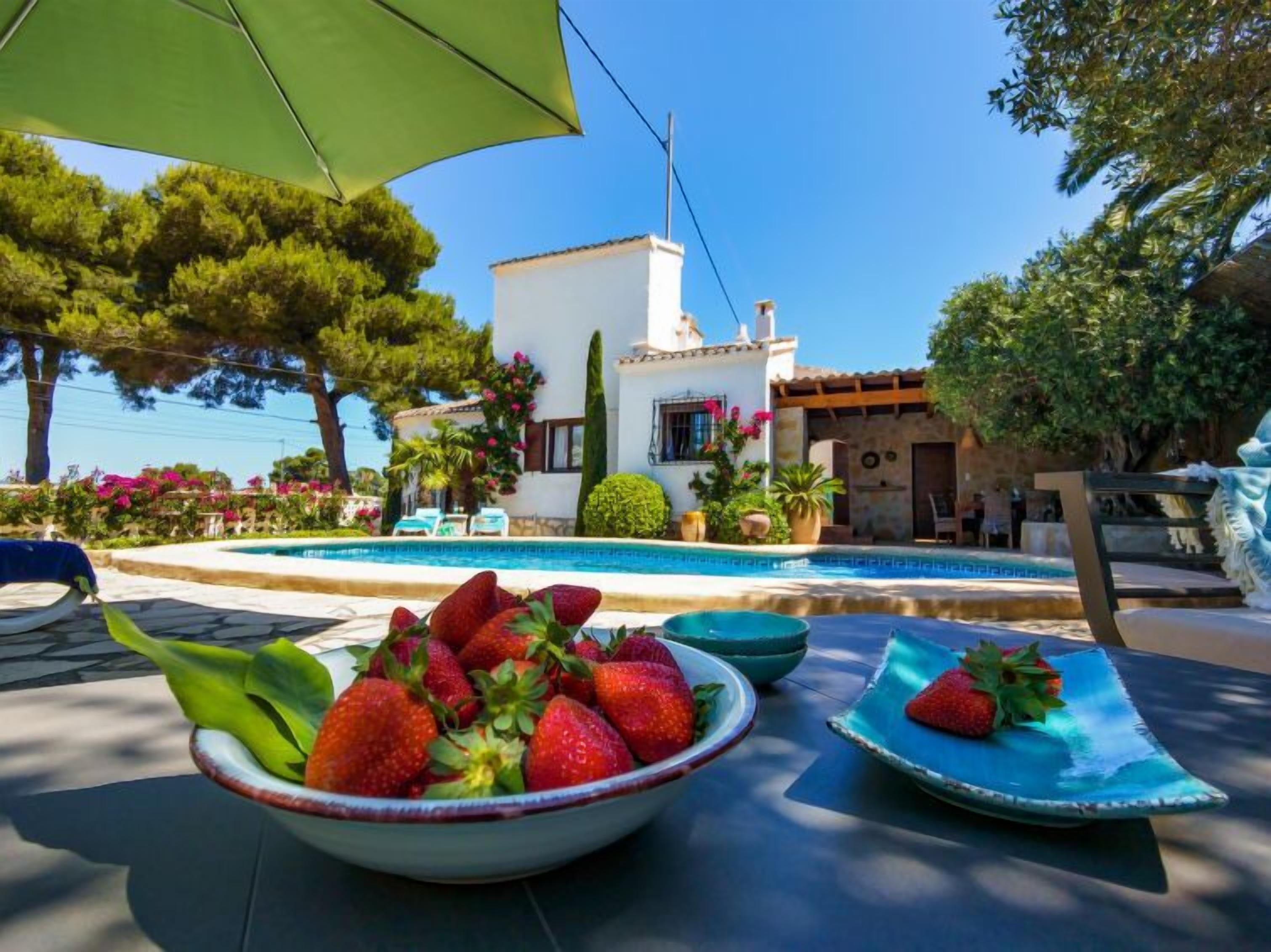A poolside area with a bowl of strawberries, a dining table, and a white building with a pool in the background.
