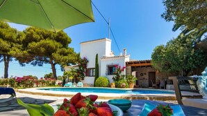 Uma área à beira da piscina com uma taça de morangos, uma mesa de jantar e um edifício branco com piscina ao fundo.