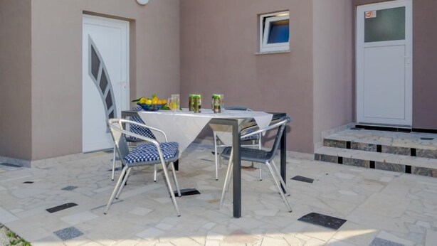 A covered outdoor dining area with a table set for two, two chairs, a bowl of fruit, and a small window. - Apartment B&B by Interhome (Medulin/Ližnjan)