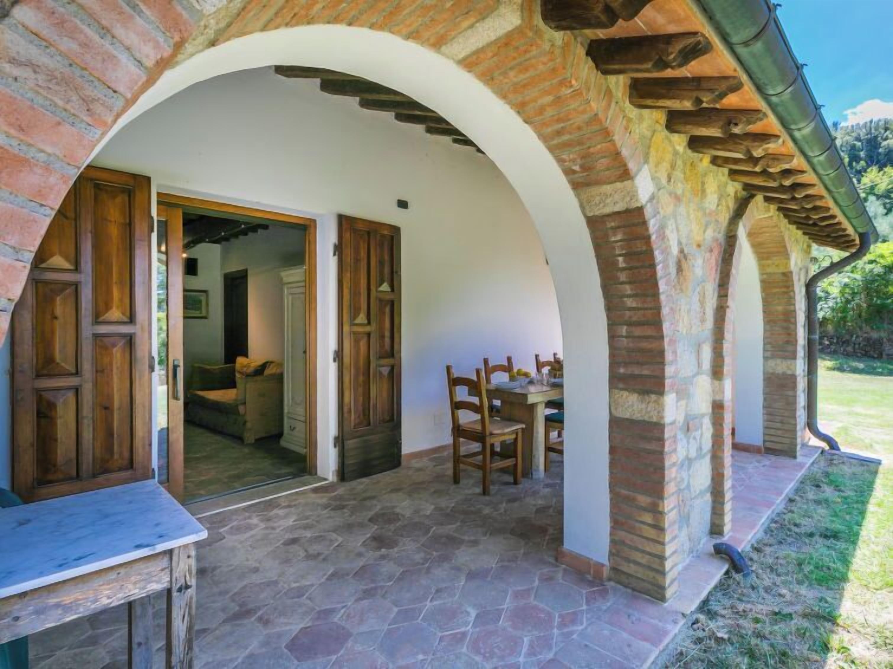 Une terrasse en pierre avec une table et des chaises, une porte ouverte donnant sur une pièce, et un mur en briques avec une porte en bois.